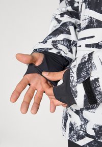 Juodai baltas raštuotas švarkas su thumb holes, pasižymintis pritaikytu dizainu, minkštos medžiagos tekstūra, reguliuojamomis rankovėmis ir velcro akcentais.