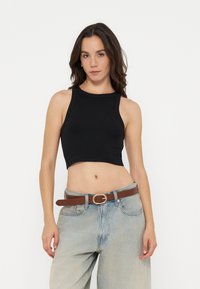 Femme portant un crop top noir sans manches associé à un jean taille haute bleu clair et une ceinture marron, debout devant un fond uni.