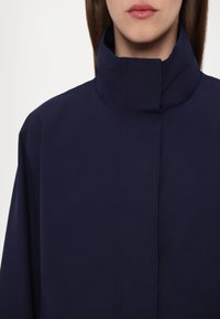 Navy blue jacket with high collar, made from smooth fabric. Features minimal stitching and two front pockets. Simple, modern design.