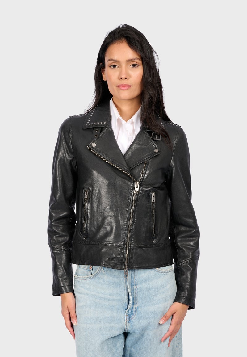 Mujer vestida con una chaqueta de motorista de cuero negra con cremalleras sobre una camisa blanca y jeans de mezclilla azules claros, de pie contra un fondo claro y liso.