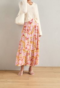 Jupe maxi florale en rose, jaune et orange avec des volants superposés, associée à un pull à col en V crémeux et duveteux et des talons beiges à lanières.