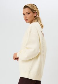 Felpa color crema con una vestibilità ampia, polsini a costine e logo stampato sul retro. Indossata con pantaloni scuri a costine.