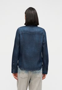 Γυναίκα με σκούρα μαλλιά μήκους ως τους ώμους, φορώντας σκούρο μπλε πουκάμισο από τζιν και ανοιχτόχρωμο τζιν, με την πλάτη προς τα εμάς, σε απλό φόντο.