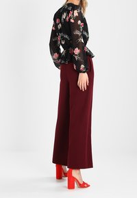 Donna in piedi che indossa una blusa nera trasparente a fiori, pantaloni larghi bordeaux a vita alta e sandali con tacco quadrato rossi.