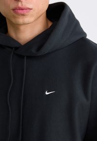 Nærbilde av en person som har på seg en svart hettegenser med en hvit Nike-svoosh-logo på brystet, som viser lepper og hake.