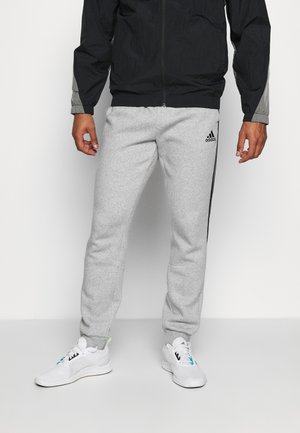 Personne portant un pantalon de survêtement gris Adidas avec des rayures noires et des baskets blanches, debout devant un fond clair uni.