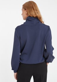 Navyblauer Pullover mit hohem Kragen, langen Ärmeln und lockerer Passform. Eine glatte Textur mit leichtem Raffung an den Bündchen.