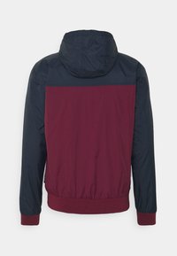 Veste à capuche avec corps bordeaux et manches, épaules et capuche bleu foncé, avec poignets et taille élastiques.