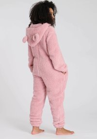 Roze, fluffy onesie met een capuchon die afgeronde oren heeft, lange mouwen en elastische manchetten. Zachte textuur en zijzakken.