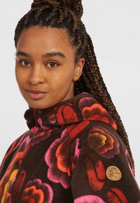 Bruine fleecejack met grote bloemmotieven in roze, rood en oranje, met een knoopsluiting en een houten logoaccent op de mouw.