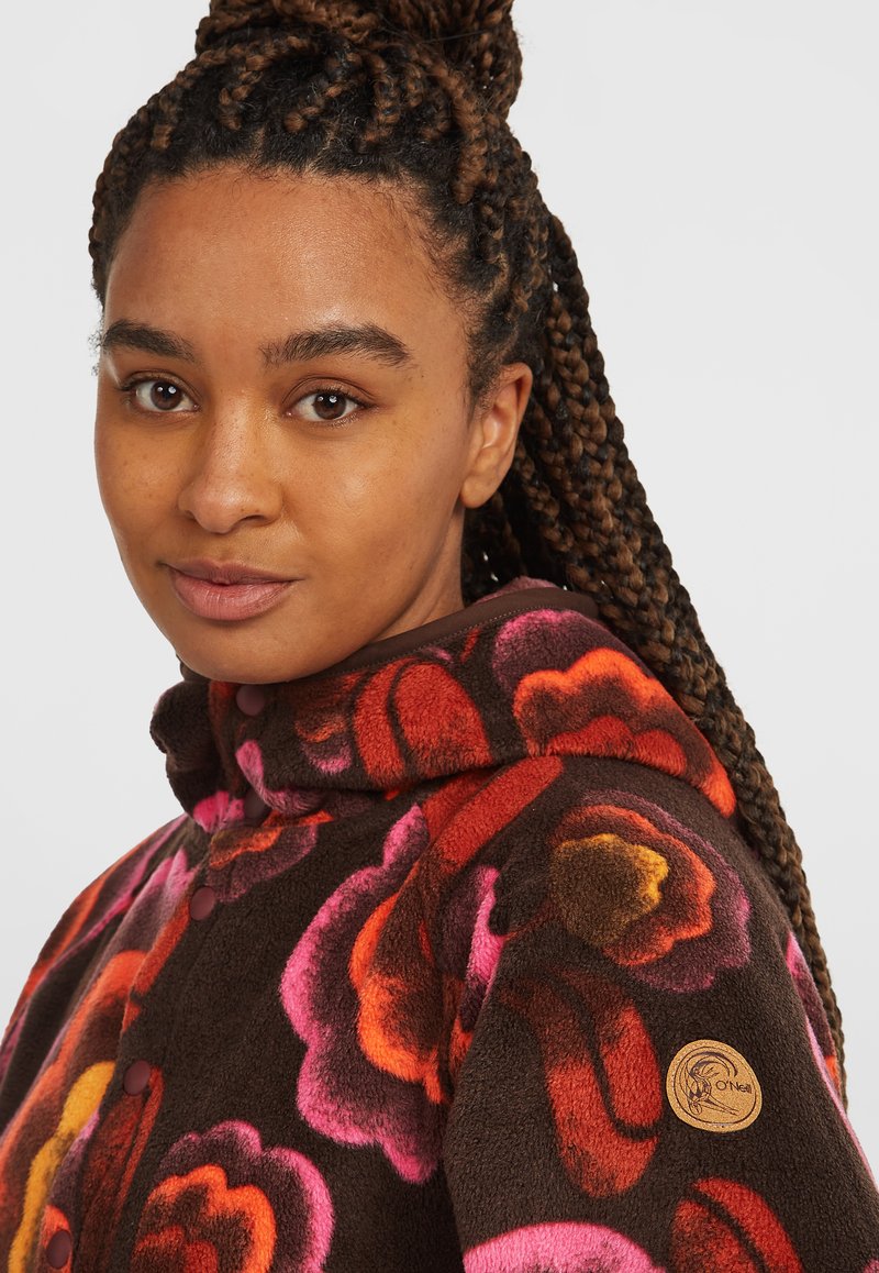 Braune Fleecejacke mit großen Blumemustern in Pink, Rot und Orange, mit Knopfverschluss und einem hölzernen Logopatch am Ärmel.