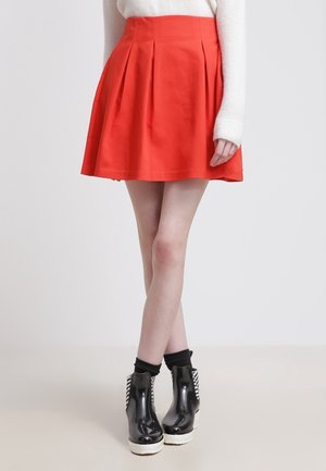 Kobieta ubrana w czerwoną, plisowaną spódnicę z wysokim stanem, zestawioną z czarno-białymi botkami na platformie oraz białą bluzką z długim rękawem.