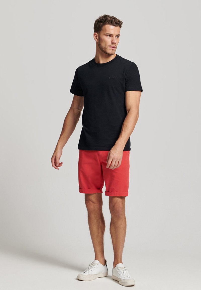 Camiseta de algodón negra combinada con shorts rojos, con dobladillos enrollados. El conjunto se completa con zapatillas blancas sobre un fondo neutro.