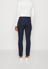 Person klädd i smala marinblå byxor, vit långärmad tröja och vita sneakers, står vänd bort med ryggen mot en enfärgad ljus bakgrund.