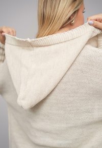 Beżowy dzianinowy hoodie z teksturowanym, luźnym materiałem. Posiada kaptur i surowe wykończenie w okolicy dekoltu.