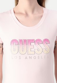 Kobieta nosząca jasnoróżową koszulkę z dużym napisem "GUESS" w różowych gradientowych literach oraz "LOS ANGELES" poniżej w małych cyrkoniach.