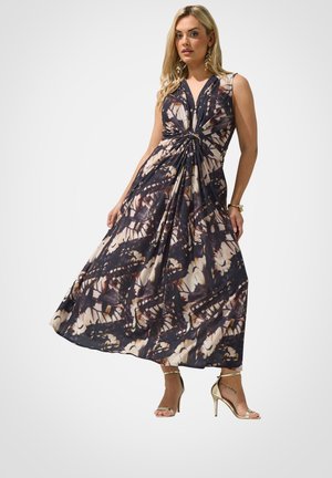 Blonde Frau trägt ärmelloses, gedrehtes Knoten-Tie-Dye-Maxikleid mit tiefem V-Ausschnitt und goldenen Knöchelriemen-Pumps, steht vor weißem Hintergrund.