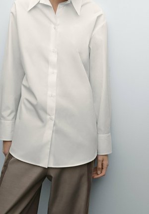 Personne portant une chemise blanche unie boutonnée à manches longues et un pantalon marron clair sur un fond clair uni.