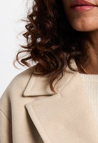 Pelo rizado castaño oscuro, abrigo de lana beige con solapa ancha, suéter de punto color crema y collar de cadena dorada en el hombro y la parte inferior del rostro de una mujer.