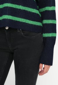 Suéter de punto a rayas en azul marino y verde con mangas largas, combinado con jeans negros ajustados y una mano parcialmente visible.