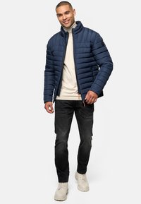 Navy blue puffer jacket with horizontal stitching, high collar, and zipper. Paired with light gray top, dark jeans, and beige boots.