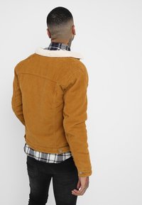 Kostymjacka i senapsgul färg med fårskinnskrage; har en figursydd design och två bakfickor, lagd över en rutig skjorta.