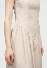 Μπεζ αμάνικο φόρεμα με εφαρμοστό μπούστο και λεπτομέρεια φούστας με πιέτες, που φοριέται από άτομο με τα χέρια χαλαρά στο πλάι.