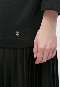 Czarny top z zaokrąglonym dołem, luźnym krojem, ozdobiony małym metalicznym logo. Ręka oparta na plisowanej czarnej spódnicy.