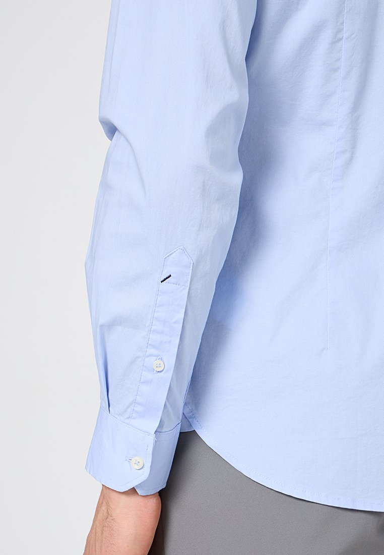 Camicia a maniche lunghe di colore azzurro chiaro con una texture liscia, polsini con bottoni e un colletto elegante. Presenta un accento nero sottile sulla manica.