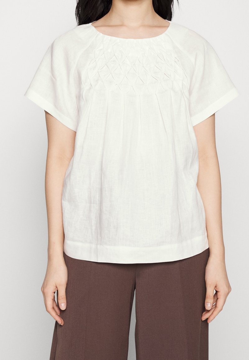 Femme portant un chemisier blanc à manches courtes avec une couture en motif losange sur la poitrine et un pantalon marron à jambes larges.