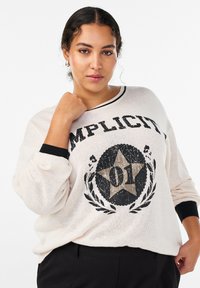 Gaiši bēšs džemperis ar melnām detaļām. Uz tā ir liels grafisks attēls ar zvaigzni un vārdu "IMPLICIT", kā arī fakturēta adījuma raksts.