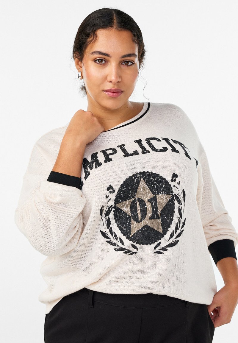 Gaiši bēšs džemperis ar melnām detaļām. Uz tā ir liels grafisks attēls ar zvaigzni un vārdu "IMPLICIT", kā arī fakturēta adījuma raksts.