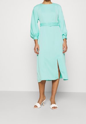 A long mint green dress with a round neck, belted waist, and puff sleeves. It features a front slit and is paired with white heeled sandals.