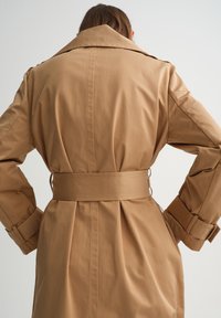 Persoon draagt een beige trenchcoat met een brede riem en manchetbanden, handen in de zij, gezien van achteren tegen een effen achtergrond.