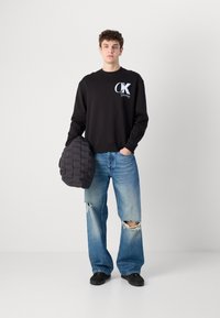 Hanorac negru cu logo alb "CK", asociat cu blugi desenați, de culoare bleu deschis. Ține în mână o geantă pufă texturată, de culoare neagră. Înc footwear casual.