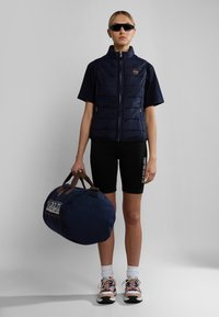 Marinblå vadderad väst med hög krage, korta ärmar och sidofickor; kombinerad med svarta cykelshorts och en marinblå duffelväska.