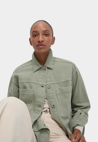 Giacca di jeans sovradimensionata verde chiaro con chiusura a bottoni, tasche sul petto e maniche arrotolate. Indossata sopra un top beige e pantaloni.