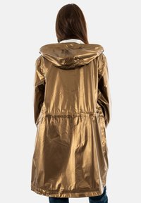 Imperméable doré métallique avec capuche, taille cintrée, manches longues et tissu léger et brillant. Présente une coupe droite et une coupe décontractée.