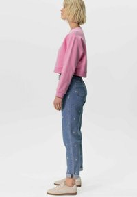 Sweatshirt rose court, jeans en denim bleu clair avec broderies florales, et baskets beiges. Cheveux coupés courts, en ondulations.