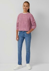 Hellrosa gestrickter Pullover mit strukturiertem Muster, kombiniert mit blauen Skinny-Jeans und weißen Sneakers, vor einem neutralen Hintergrund stehend.