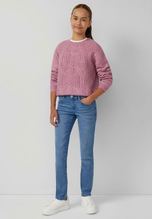 Lichtroze gebreid sweater met een gestructureerd patroon, gecombineerd met blauwe skinny jeans en witte sneakers, staande tegen een neutrale achtergrond.
