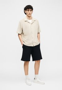 Joven de pie con las manos en los bolsillos, vistiendo una camisa beige de manga corta abotonada, pantalones cortos negros, calcetines blancos y zapatillas blancas.