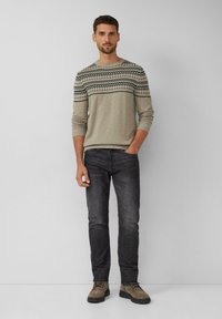 Man staat met één hand in zijn broekzak, draagt een beige gebreide trui, zwarte jeans en bruine veterschoenen tegen een effen achtergrond.