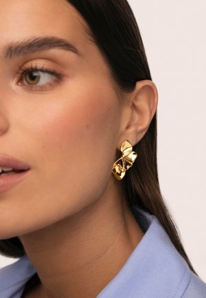 Pendiente llamativo en tono dorado con un diseño orgánico y escultórico. Superficie lisa y brillante con una forma retorcida, llevado en la oreja izquierda.