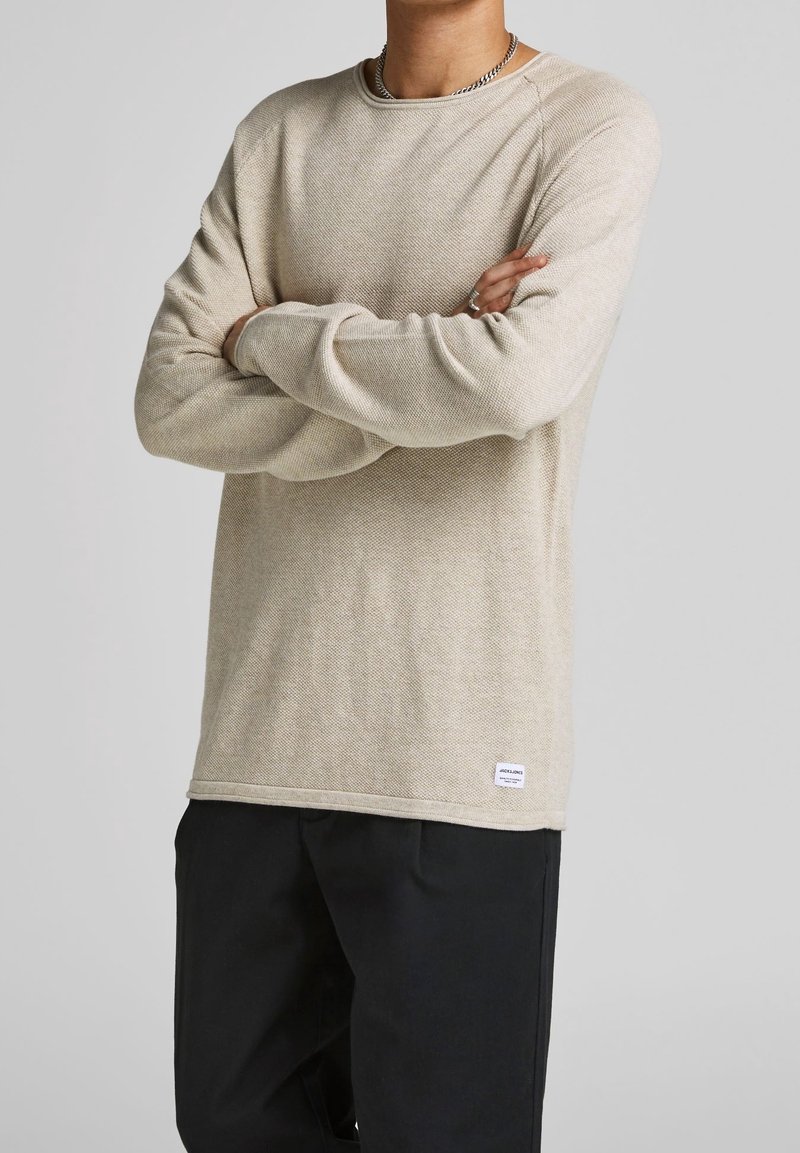 Chemise beige à manches longues en matériau texturé, avec un col rond et une petite étiquette de marque en bas à gauche. Associée à un pantalon noir.