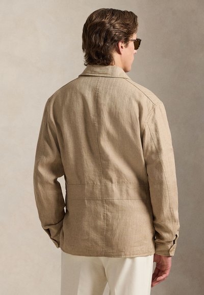 Man wearing light brown linen jacket and white pants, shown from the back against a neutral beige background.