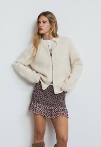 Jersey de color crema con cremallera y mangas raglán, combinado con una mini falda burdeos estampada con dobladillo con volantes.