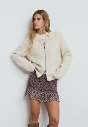 Jersey de color crema con cremallera y mangas raglán, combinado con una mini falda burdeos estampada con dobladillo con volantes.