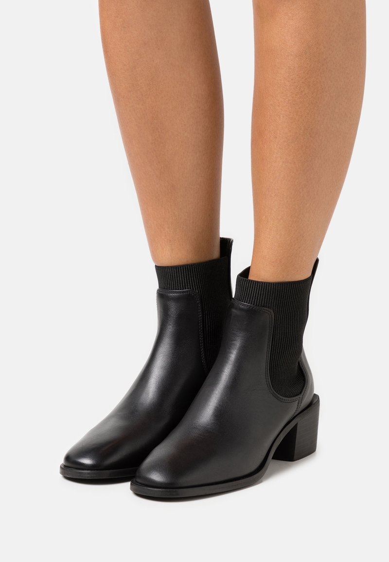 Bottines en cuir noir avec accents côtelés style chaussette, bout pointu et talon bloc bas, présentant une texture lisse et un design épuré.