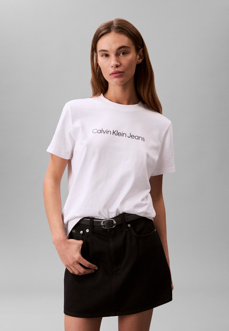 Camiseta blanca de algodón con la inscripción "Calvin Klein Jeans" en negro, combinada con una falda vaquera negra y un cinturón negro, sobre un fondo gris.